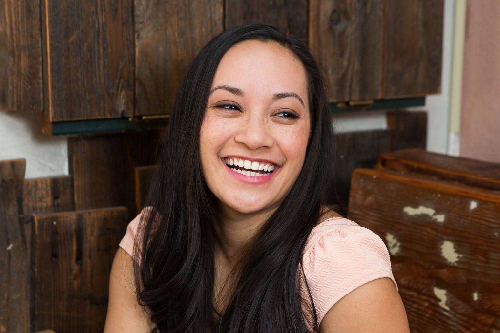 Dark-haired woman with bright white teeth & pink shirt laughing at dentist